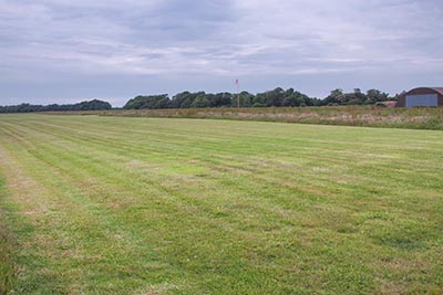 The site of Lymington Airfield with a grass runway in the foreground - that is still in use today - and a Second World War, Blister hanger visible on the right